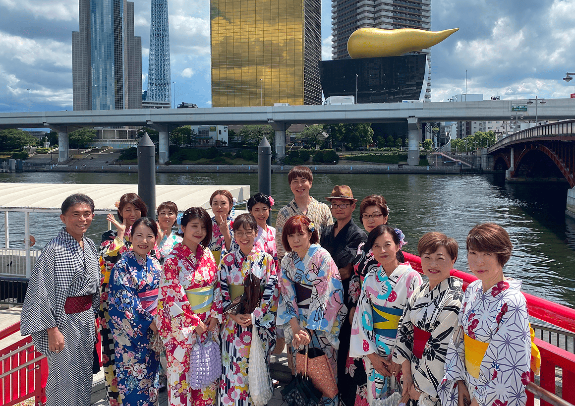 東京・隅田川沿いで、浴衣姿のメンバーが笑顔で記念撮影。背景にはスカイツリーとアサヒビール本社ビル、川には夏空が広がる。
